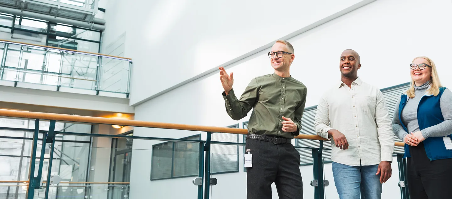 Kolme hymyilevää henkilöä seisovat vierekkäin valoisalla toimistorakennuksen käytävällä ja katsovat kauas.