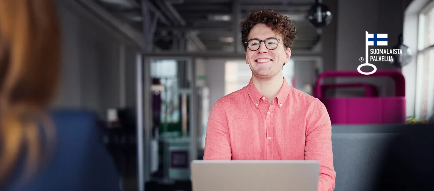 Leveästi hymyilevä miesoletettu istuu neuvottelupöydän ja tietokoneen ääressä toista henkilöä vinosti vastapäätä..