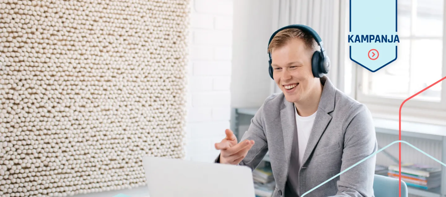 Henkilö kokouspöydän ja kannettavan tietokoneen äärellä katsoo hymyillen ruutuun ja elehtii kädellään. 