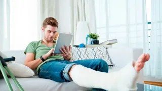 Henkilö, jolla on kipsattuna jalka, istuu sohvalla tabletti-tietokone kädessä.