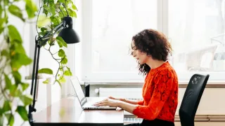 Nainen istuu kirjoituspöydän ääressä ja kirjoittaa tietokoneella - viherkasvi etualalla.
