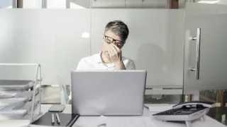 Henkilö istuu kannettavan tietokoneen ääressä ja hieroo otsaansa.