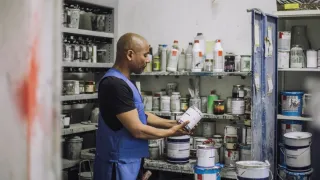 Työntekijä katselee erilaisia kemikaaleja täynnä olevassa varastossa kädessään olevaa maalipurkkia.