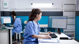 Sairaanhoitaja istuu pöydän ääressä ja tarkastelee tietokoneen ruutua.
