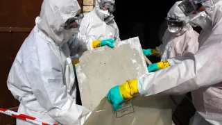 Neljä suojavarusteista asbestipurkutyöntekijää laittaa asbestia sisältävää levyä jätesäkkiin.
