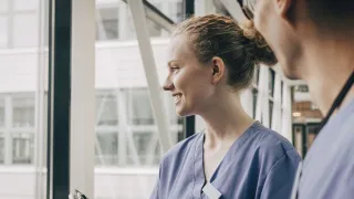 Kaksi hoitajaa katselee sairaalan ikkunasta hymyillen.