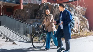 Kaksi henkilöä kävelee kadulla ja keskustelee, toinen taluttaa pyörää