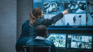 Turvallisuusalan työntekijät katsovat monitorointiruutua valvomossa. Toinen työntekijöistä osoittaa sormella ruutua. 