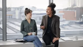 Työkaverit juttelevat keskenään istuen ikkunalaudalla