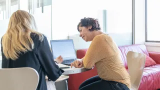 Kaksi henkiloä istuu vierekkäin työpöydän ääressä, toinen hymyilee.