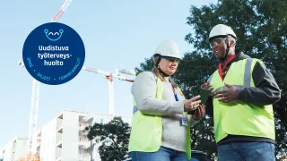 Kaksi henkilönsuojaimiin ja huomioliiveihin pukeutunutta henkilöä keskustelevat ulkona rakennustyömaa taustalla.