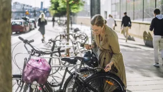 Naisoletettu henkilö avaa pyörän lukkoa kännykkä ja tavaroita käsissään kuulokkeet korvilla.