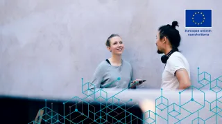 Kaksi henkilöä keskustelee kasvokkain seinän vierustalla, etualalla on tietokoneen näyttö.