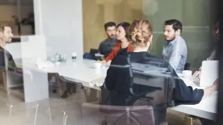 Ihmisiä istuu lasin takana neuvotteluhuoneessa. 