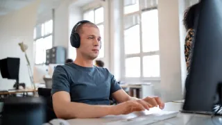 Henkilö istuu tietokoneen äärellä vastamelukuulokkeet korvillaan avokonttorissa.