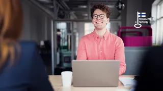 Leveästi hymyilevä miesoletettu istuu neuvottelupöydän ja tietokoneen ääressä toista henkilöä vinosti vastapäätä..