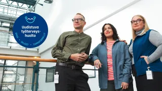 Kolme henkilöä seisovat vierekkäin valoisalla toimistorakennuksen käytävällä ja katsovat kauas.