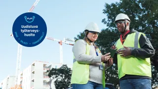 Kaksi henkilönsuojaimiin pukeutunutta henkilöä seisoo kadulla keskustelemassa, taustalla rakennustyömaa. 