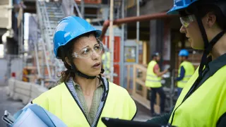 Kaksi kypäräpäistä henkilöä keskustelee rakennustyömaalla. 