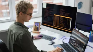 Nuori mies istuu toimistoympäristössä ja katelee yhtä kolmesta näytöstä pöydällä.