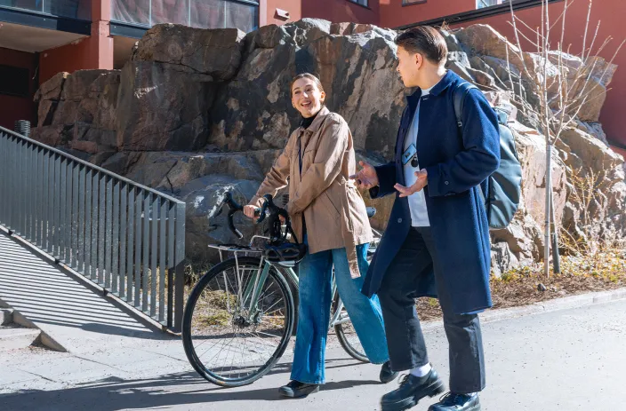 Kaksi henkilöä kävelee kadulla ja keskustelee, toinen taluttaa pyörää