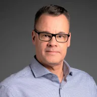 Mies silmälasit ja vaaleansininen paita päällä katsoo kohti kameraa