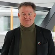 Mies musta bleiseri ja tummanvihreä korkeakauluksinen paita päällä katsoo kameraan